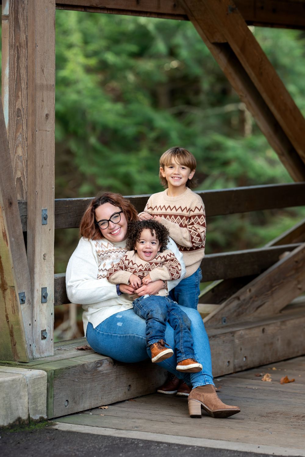 Mother sitting on covered bridge steps with two children in matching fair isle sweaters, smiling at camera in autumn.