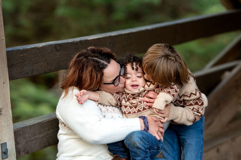 A mother hugs and kisses her two young children on a wooden bridge surrounded by green trees.