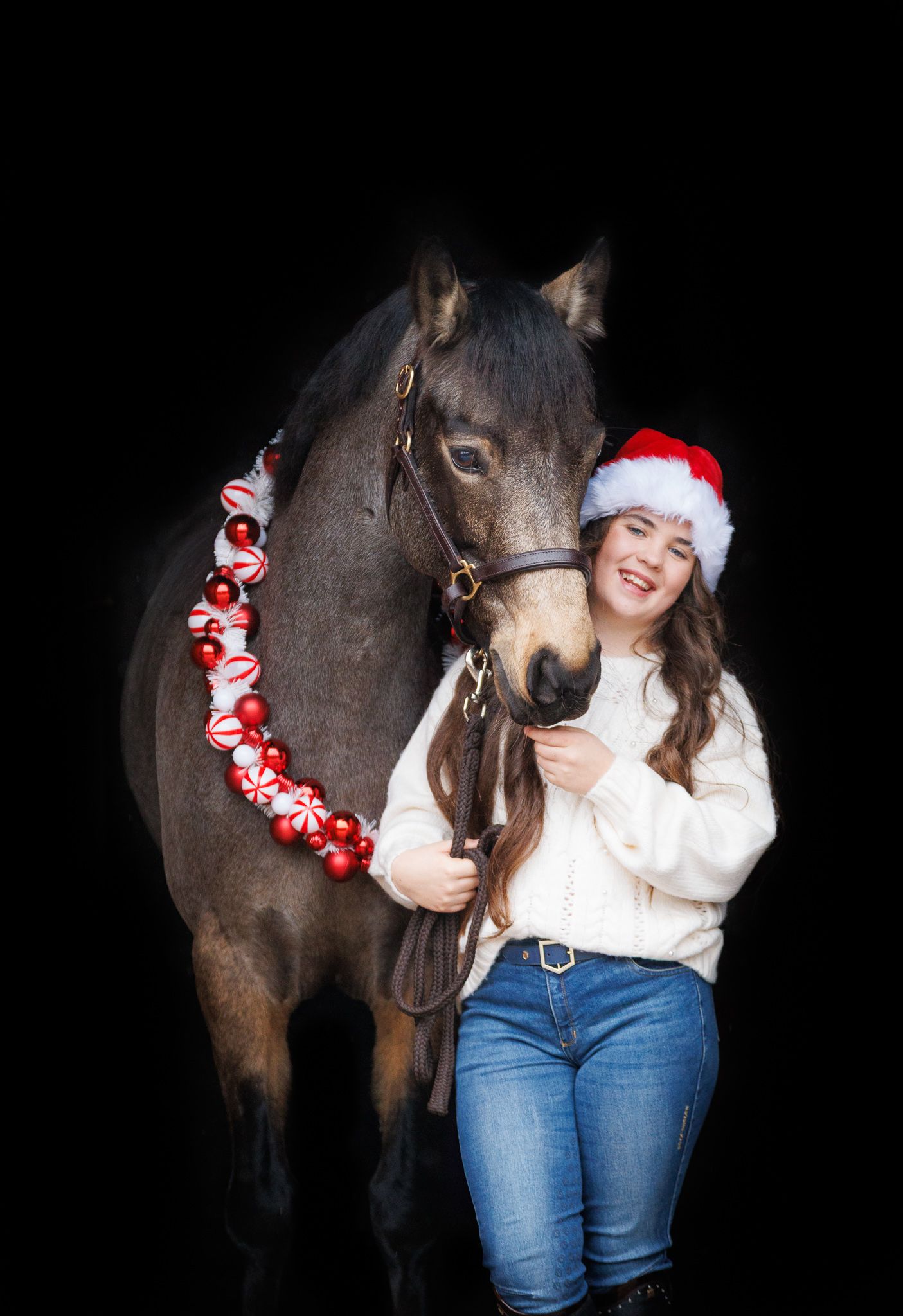A dark horse wearing festive Christmas decorations stands next to someone in jeans and a white sweater against a black background.