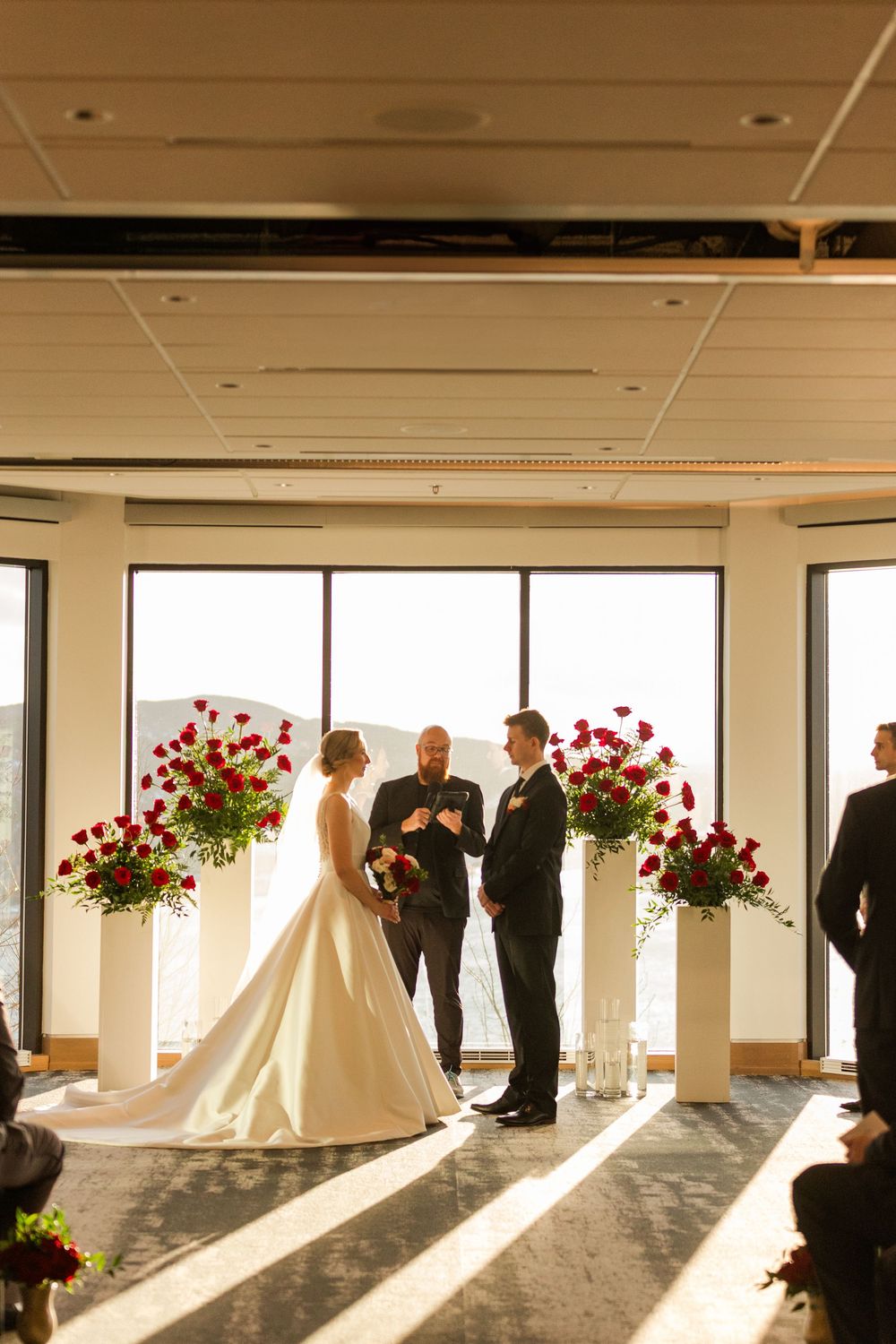 A wedding ceremony takes place in a bright sunlit room with large windows and red floral arrangements.
