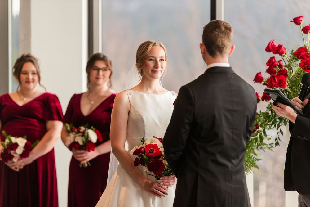 Wedding party members in burgundy dresses and black suits stand in line holding vibrant red bouquets during ceremony.