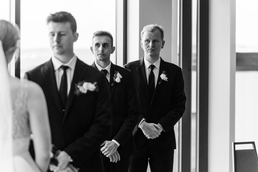 A black and white wedding photo showing groomsmen in formal attire standing in a row at a venue entrance.