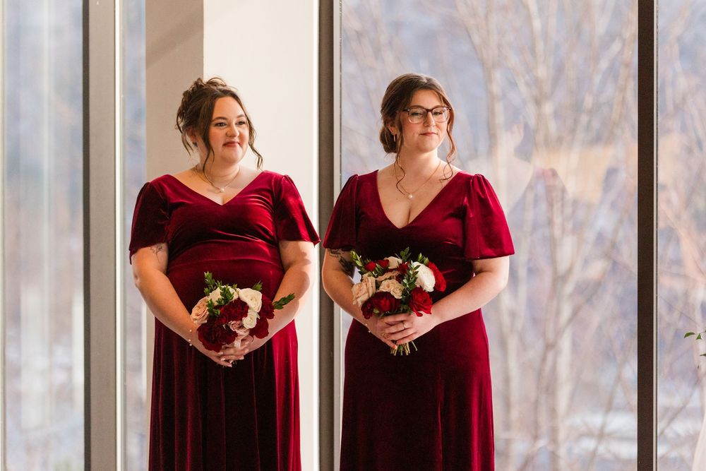 Two bridesmaids in burgundy velvet dresses hold bouquets while standing by large windows in a bright room.