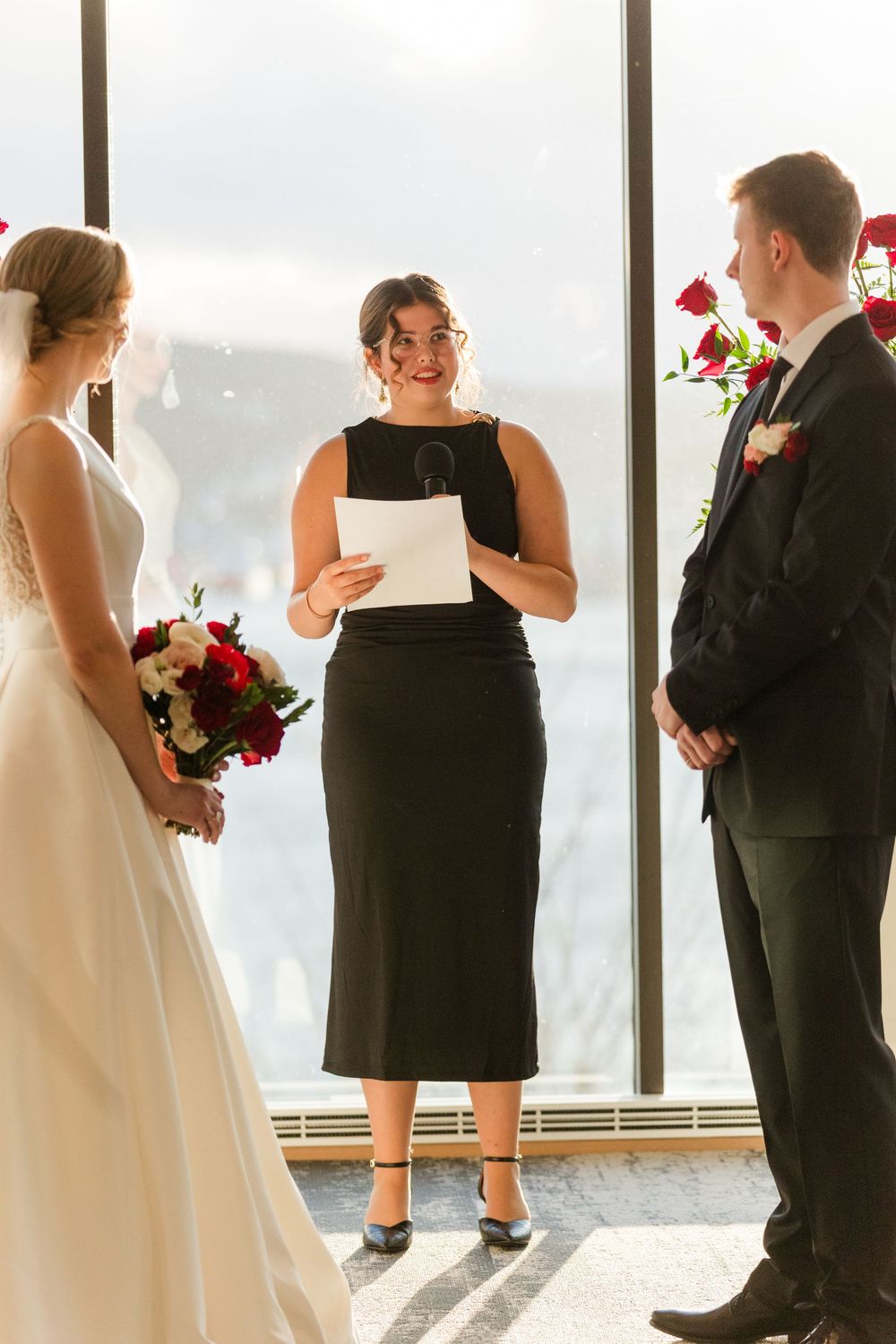 An indoor wedding ceremony with a couple and officiant standing before large windows with scenic views.