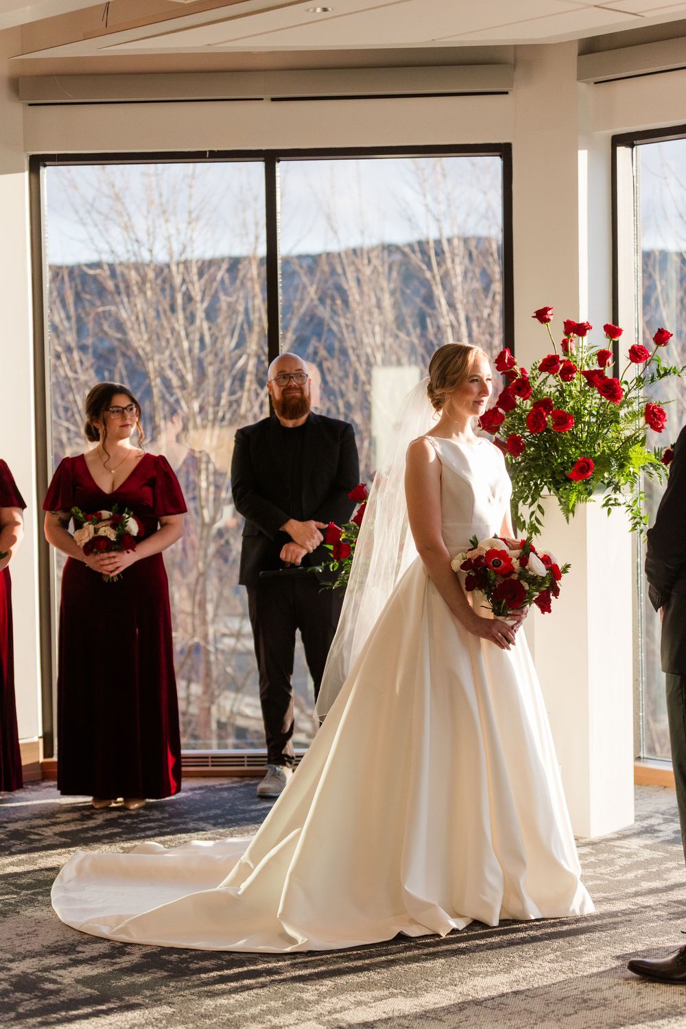 Wedding party members in burgundy and black attire stand in a sunlit venue with red rose bouquets and winter views.