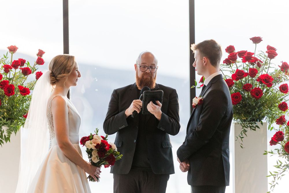 A wedding ceremony takes place in front of large windows with red floral arrangements on pedestals.
