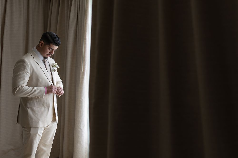 Groom in a white suit checks his phone while getting ready in a sunlit room.