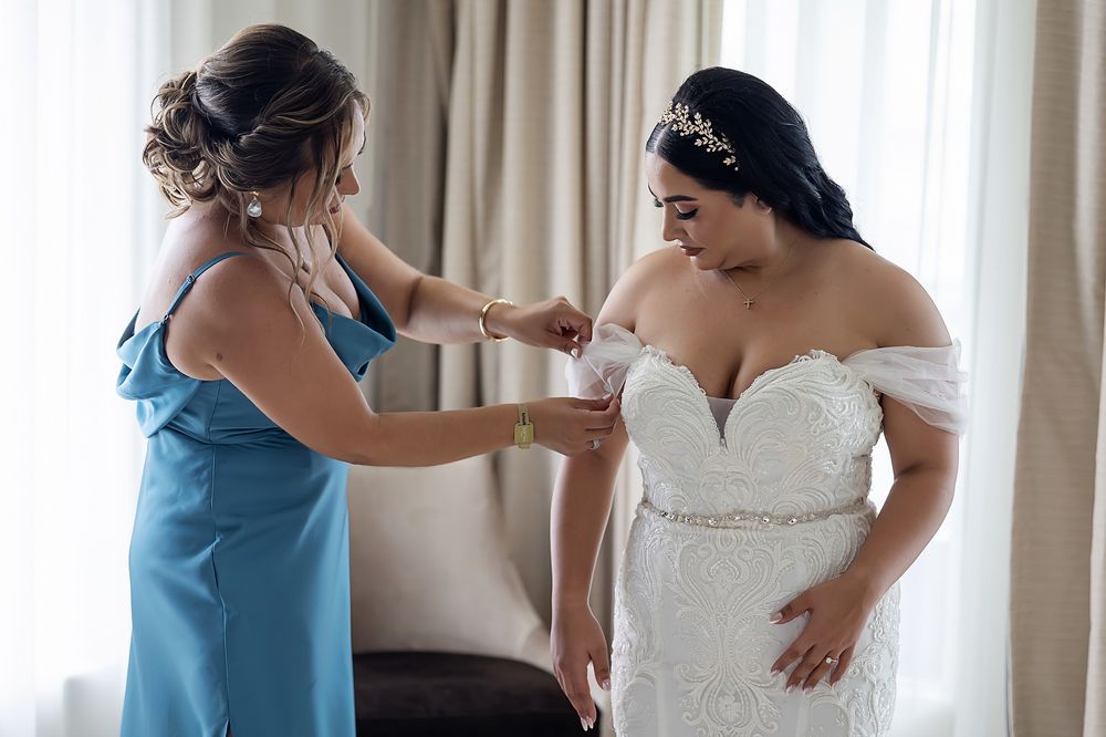 Wedding party helps bride with final dress adjustments in front of curtained window.