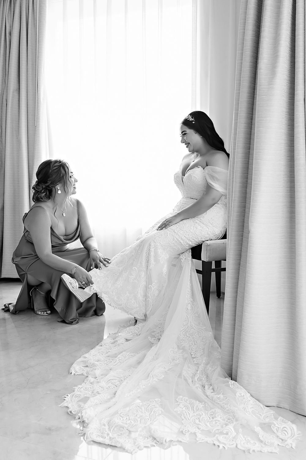 A bride in a white lace wedding gown gets help with final adjustments in a sunlit room with white curtains.