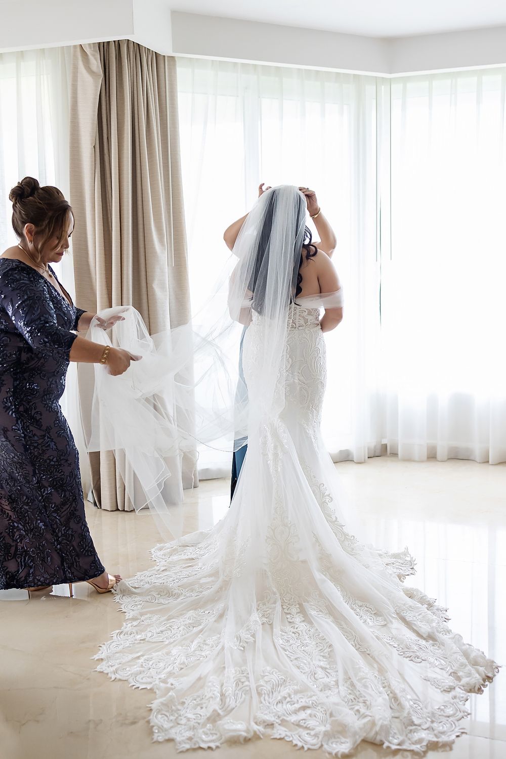 A wedding dress with intricate lace details being arranged on the floor in a bright room with sheer curtains.