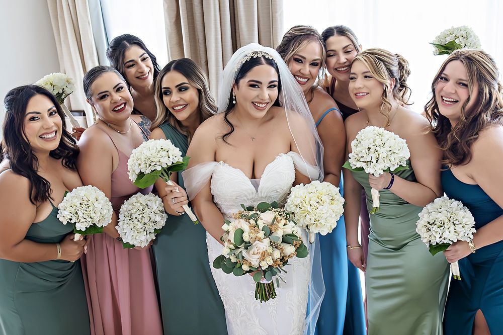 A group of bridesmaids in varying shades of blue and pink dresses pose together holding white bouquets.