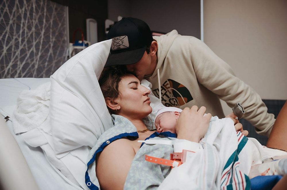 A tender moment in a hospital room as a new parent leans over to kiss their newborn baby in a hospital bed.