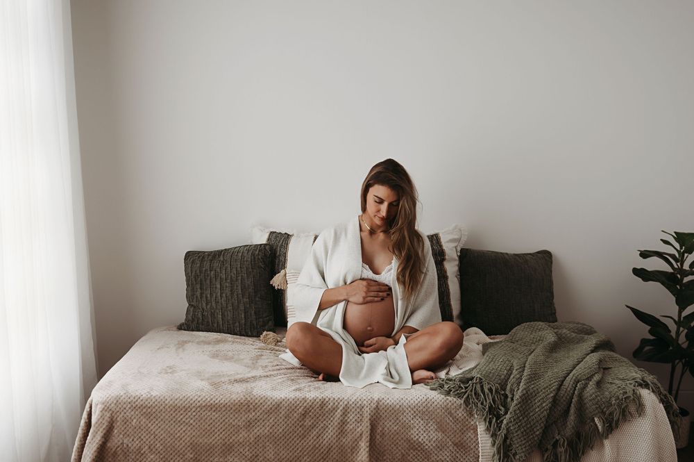 Woman in white loungewear relaxes on cozy bed with grey bedding and natural light streaming through windows.