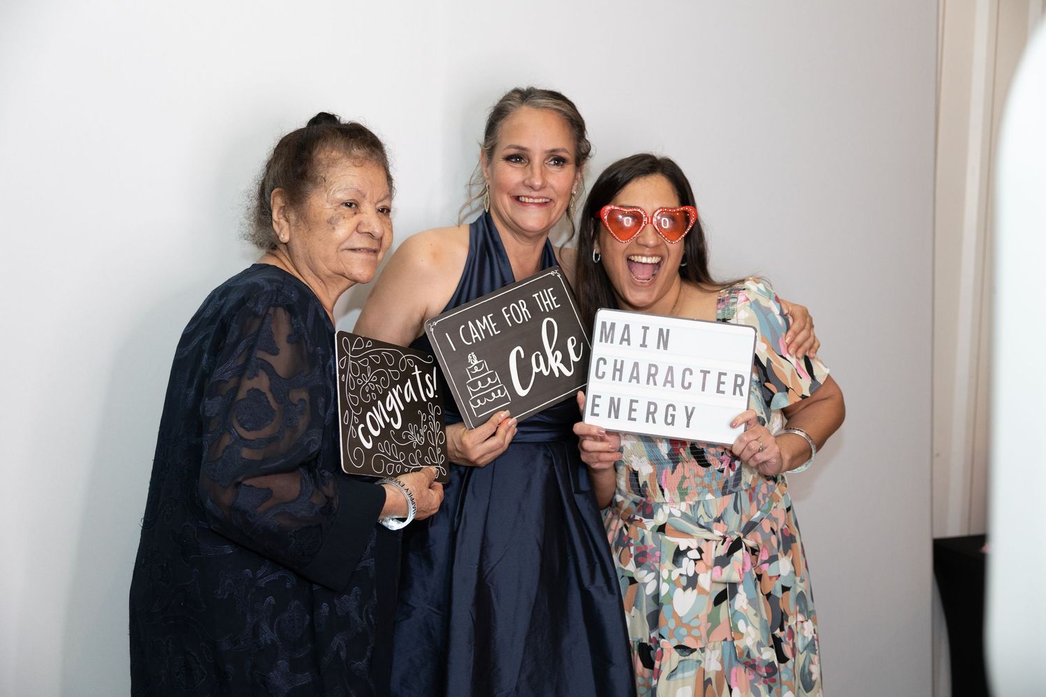 Three women posing with fun signs at a wedding photo booth celebration.