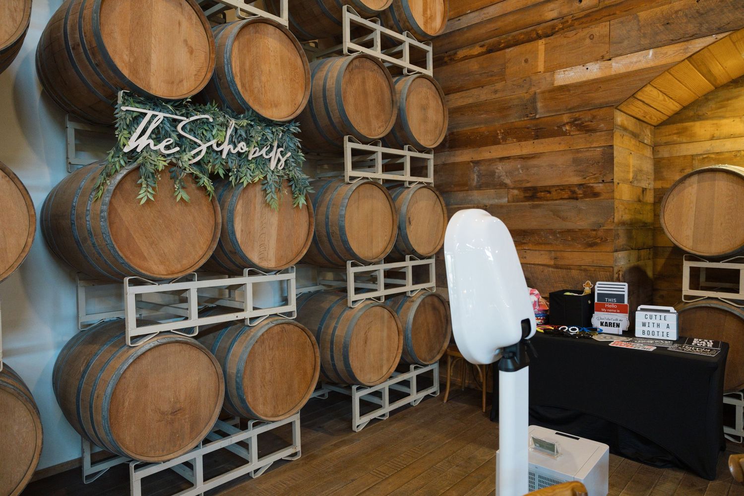 Wedding photo booth setup beside stacked oak wine barrels with a floral surname sign at a rustic winery venue.