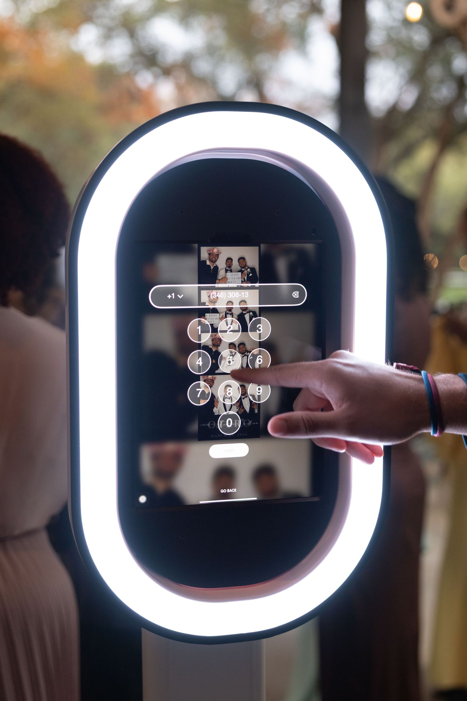 Guest using touchscreen interface on a modern oval mirror photo booth with glowing ring light at an event.
