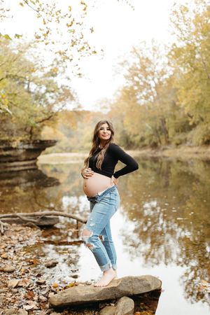 A maternity photo session by a tranquil pond with autumn leaves scattered on rocks in golden afternoon light.