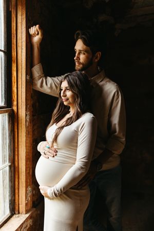 A couple stands by a rustic window in warm light, with a pregnant woman in a white dress being embraced from behind.