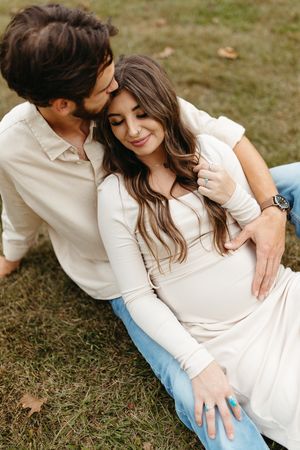 Romantic couple in white shirts and jeans sitting on grass during outdoor photo session.