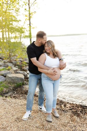 A couple embraces on a rocky beach shoreline during sunset with trees and water in the background.