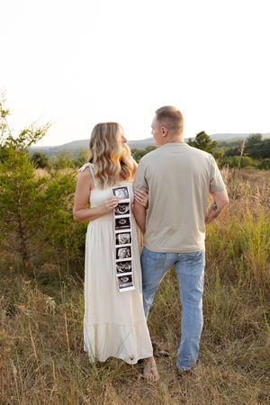 A couple stands in a grassy field holding ultrasound photos during a pregnancy announcement photoshoot at sunset.