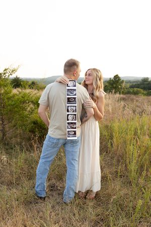 A couple stands in a sunlit meadow sharing an intimate moment while holding ultrasound photos.