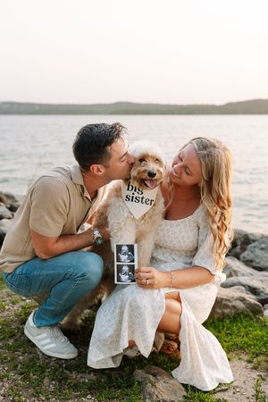 Couple on rocky shoreline at sunset with small dog wearing 'big sister' sign and ultrasound photo for pregnancy announcement.