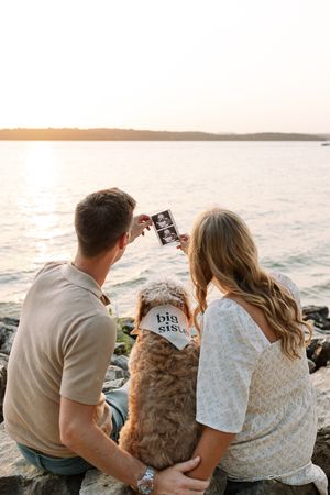 A couple and their dog sit together watching the sunset over the ocean from a rocky coastline.