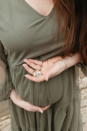 A pregnant woman in an olive green dress holds her belly while wearing delicate rings on her hand.
