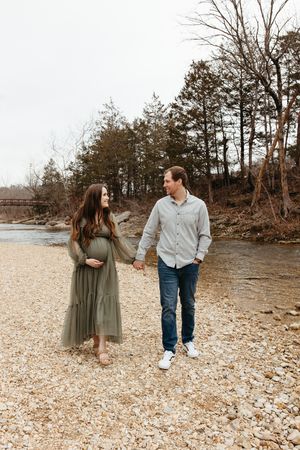 A couple shares playful moments while walking along a rocky shoreline with trees in the background.