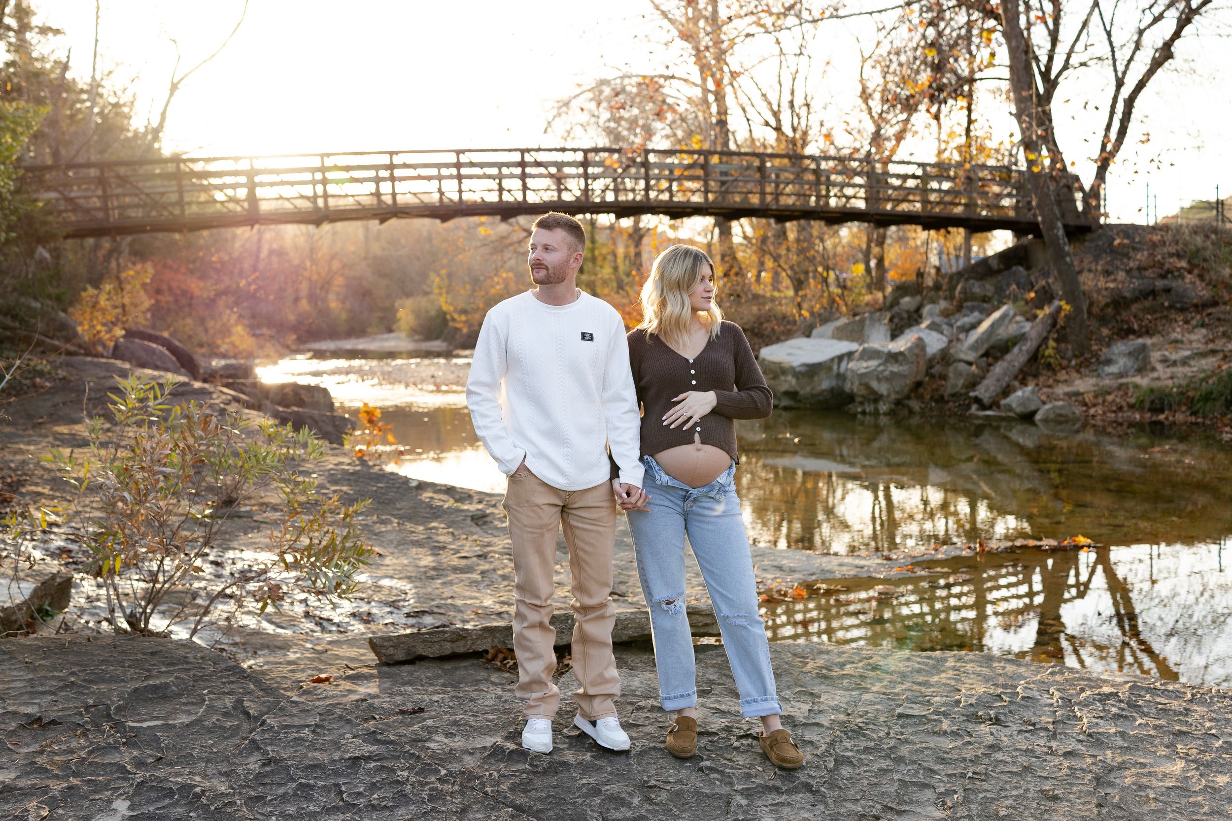 A couple walks hand in hand along a scenic wooden footbridge over a tranquil stream in an autumn setting.