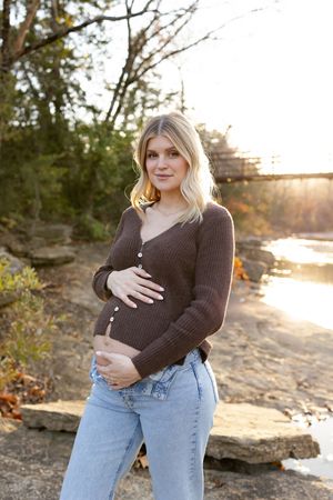 A maternity photo taken at sunset by a river with rocks and trees in the background.