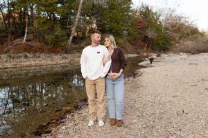 A couple stands together on a rocky beach shoreline surrounded by autumn trees and water.