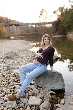 A scenic maternity portrait by a river with a stone bridge visible in the autumn landscape.