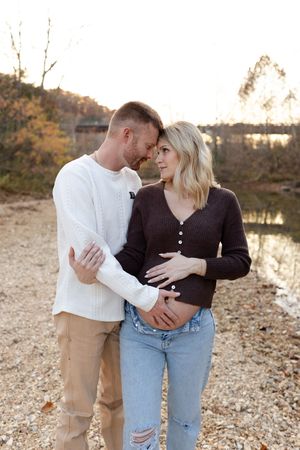 A couple in casual sweaters embrace by a lake at sunset on a rocky shore during autumn.