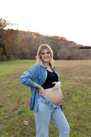 A pregnant woman in denim jacket and jeans poses in a sunny autumn field during a maternity photoshoot.