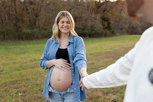 An expectant mother in a denim shirt and jeans smiles while standing in a grassy autumn field.