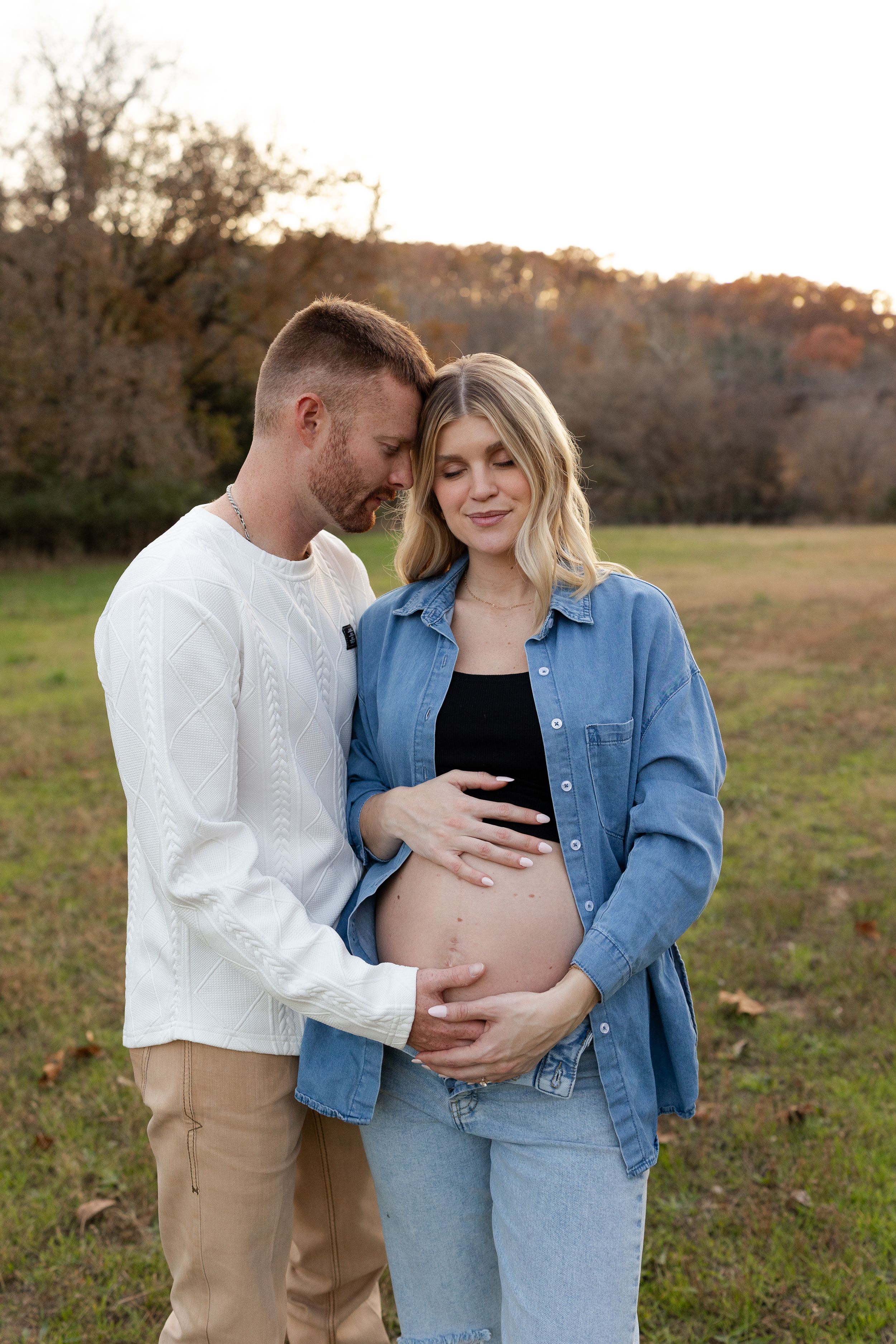 Expecting couple shares tender moments in a field at sunset while wearing denim jacket and white sweater.