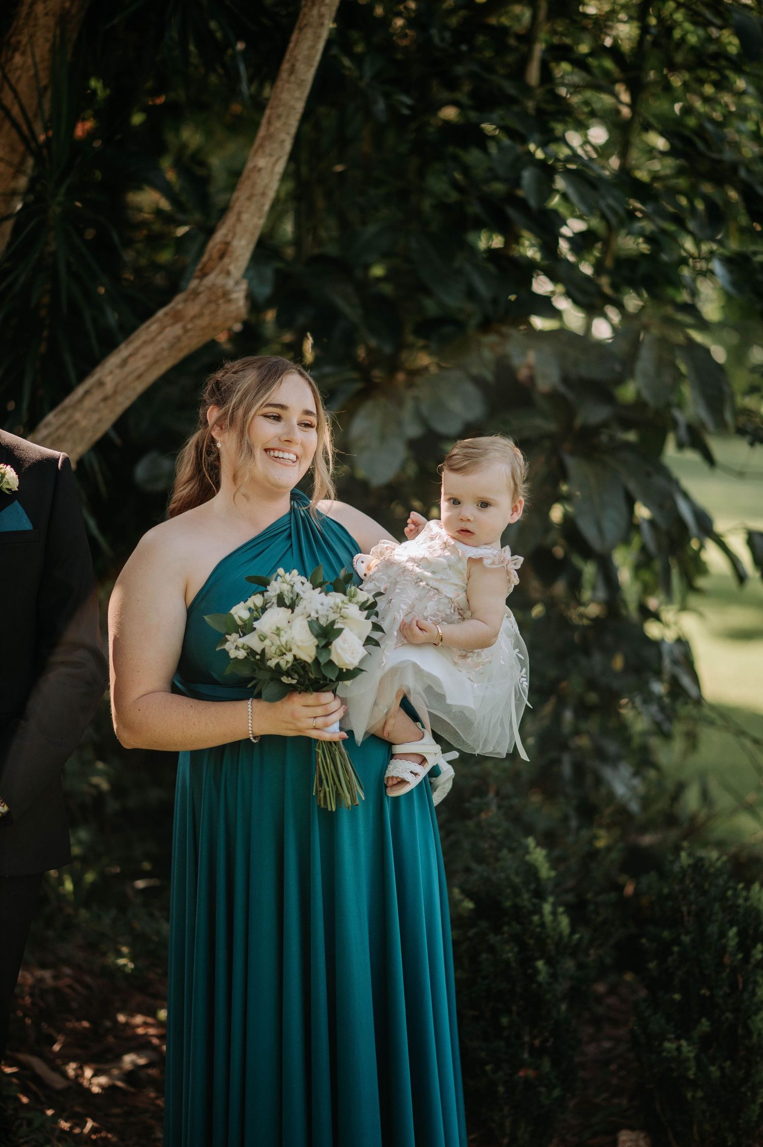 A tender moment captured under a tree as someone in a teal bridesmaid dress holds a small child and white bouquet.