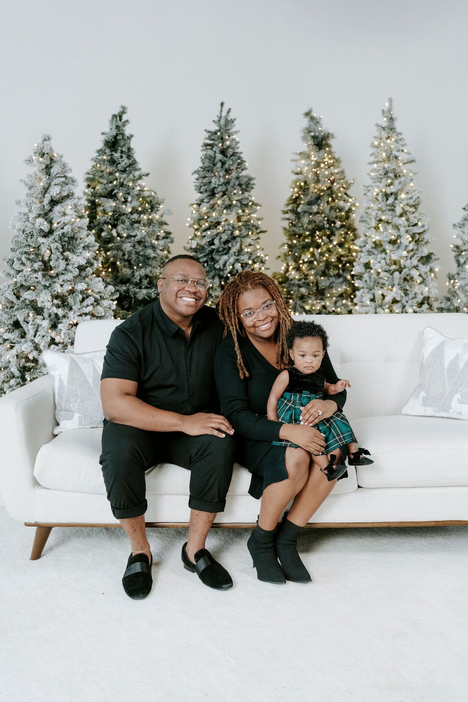 A family sits together on a white couch with snow-covered Christmas trees in the background for a holiday photo shoot.