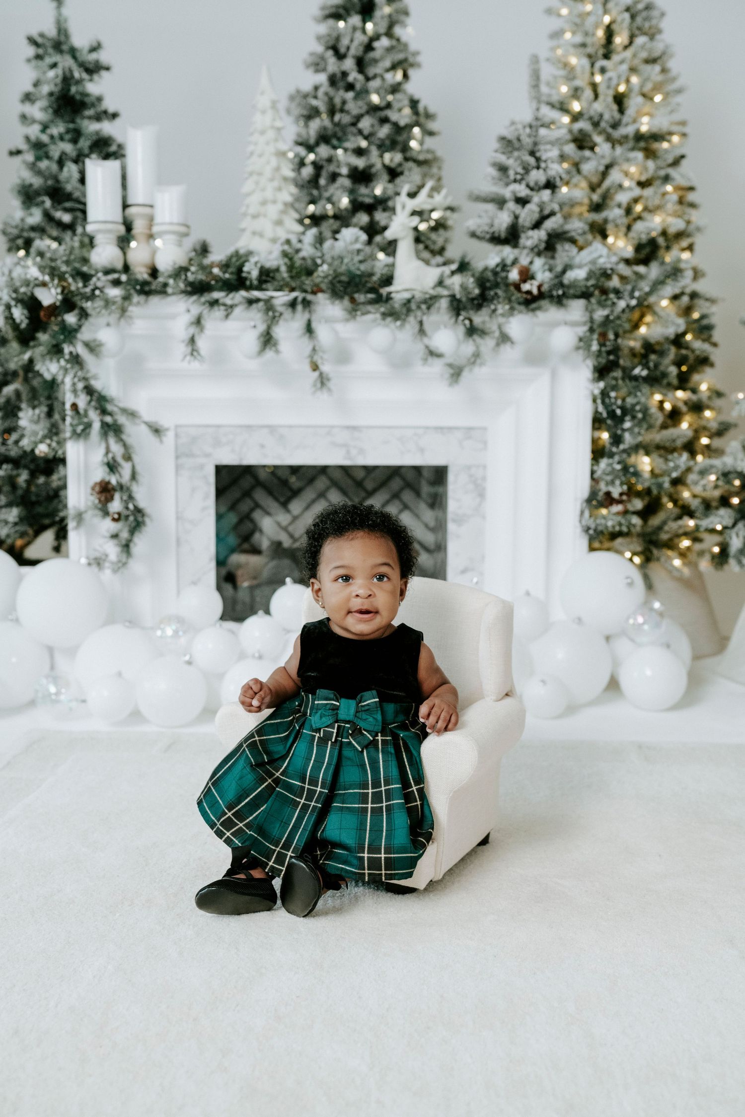 A festive holiday scene with a small child in a green plaid dress sitting in a white chair by decorated Christmas trees.