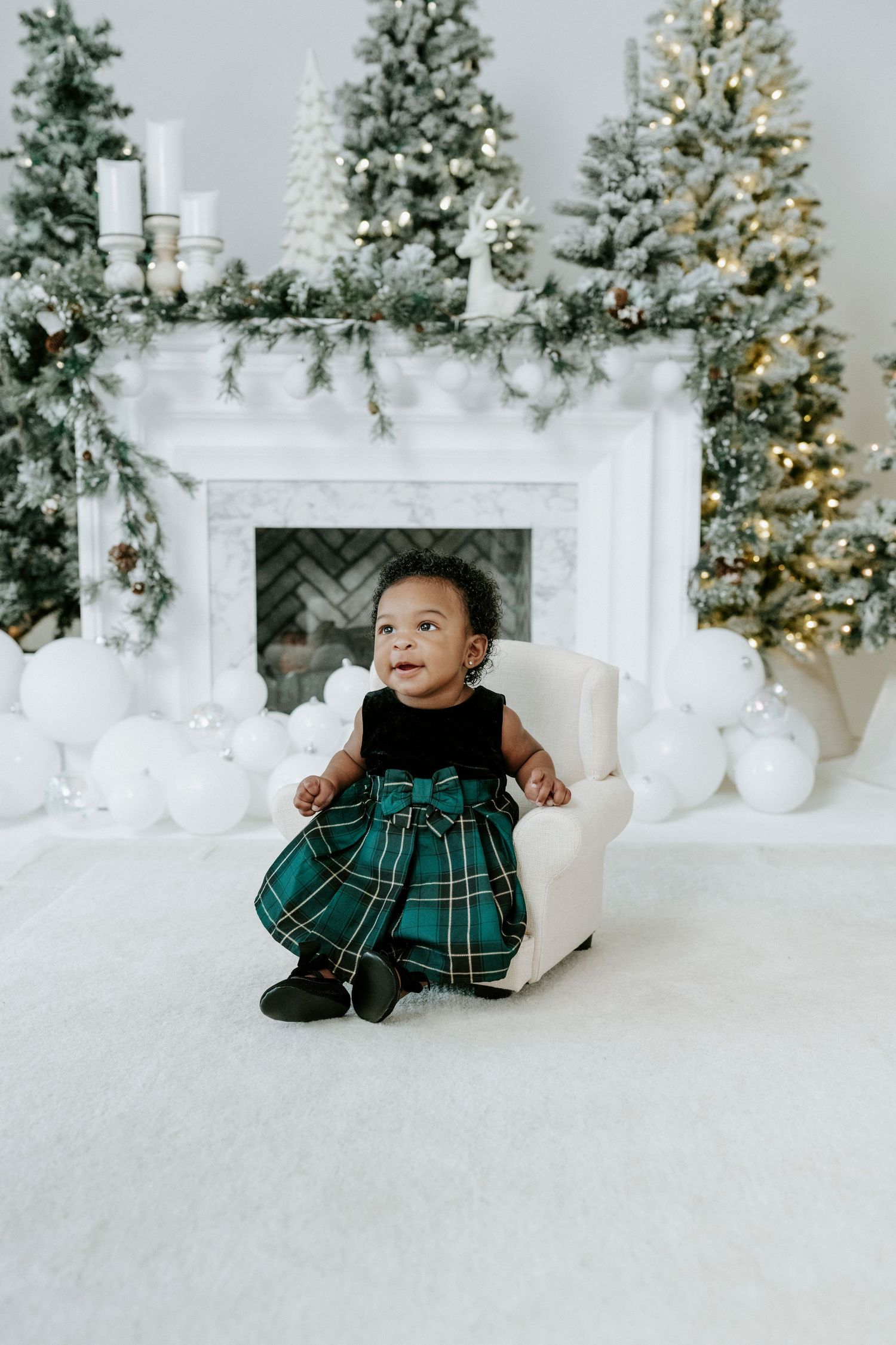 Baby in green plaid holiday dress sits on white floor with Christmas trees and decorations in background.