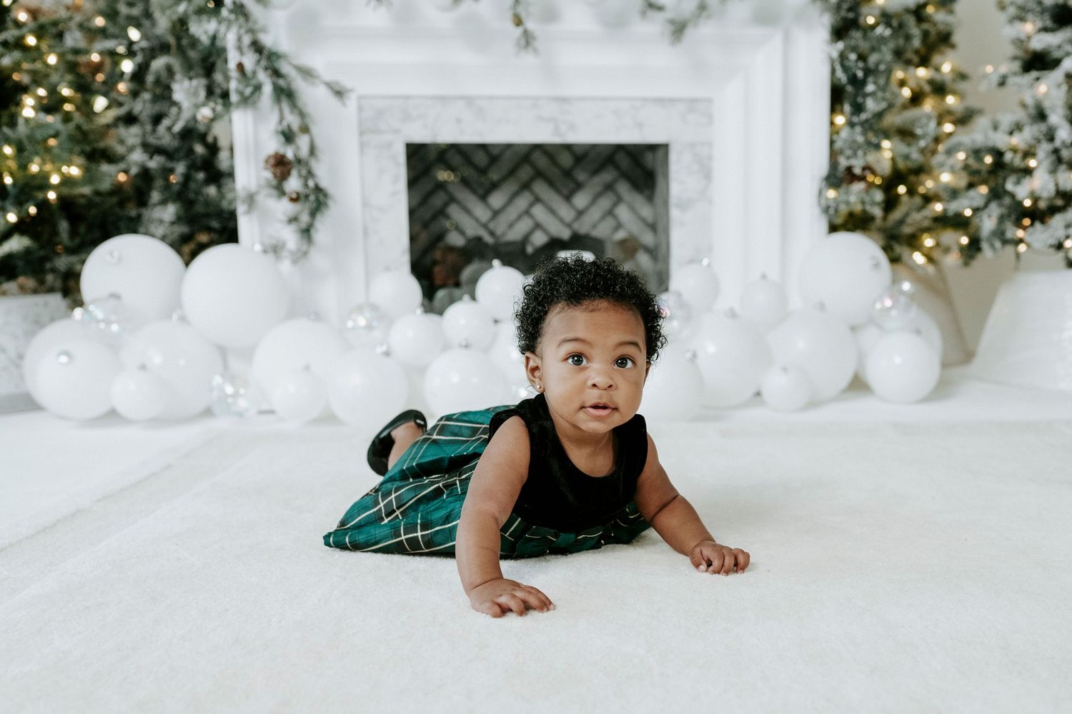 A baby in a green plaid outfit crawls on a white floor with festive holiday decorations in the background.