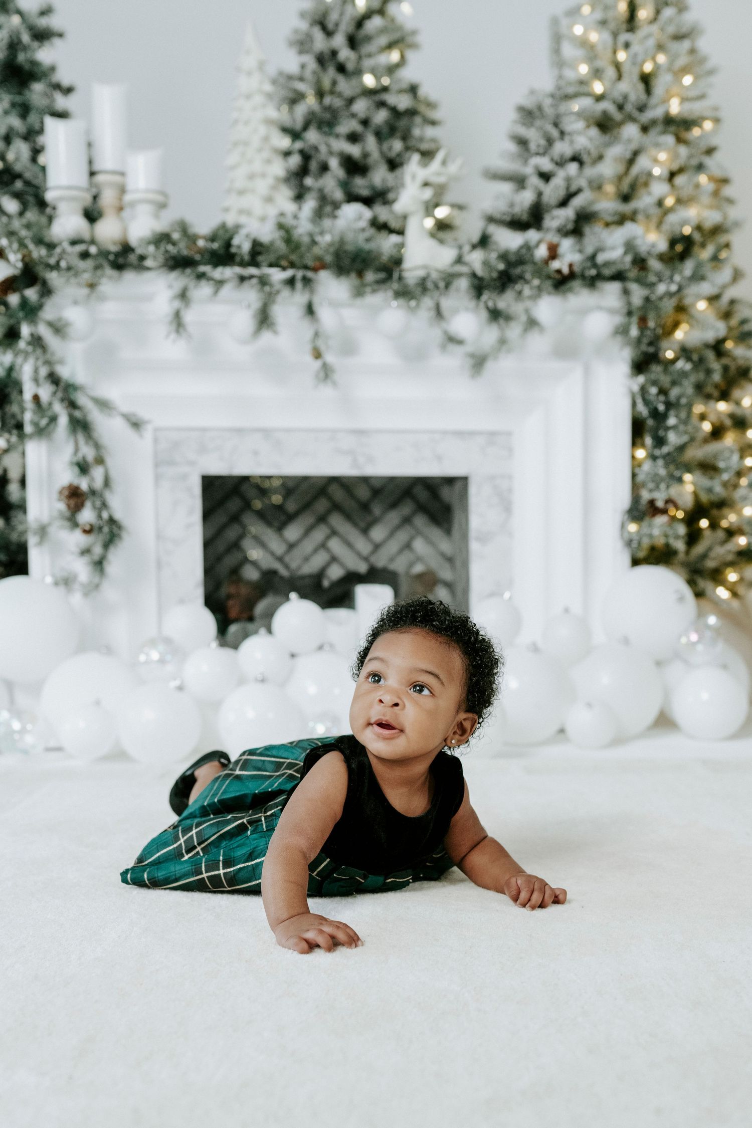 Baby in green velvet holiday dress crawls on white floor near decorated Christmas trees and fireplace.