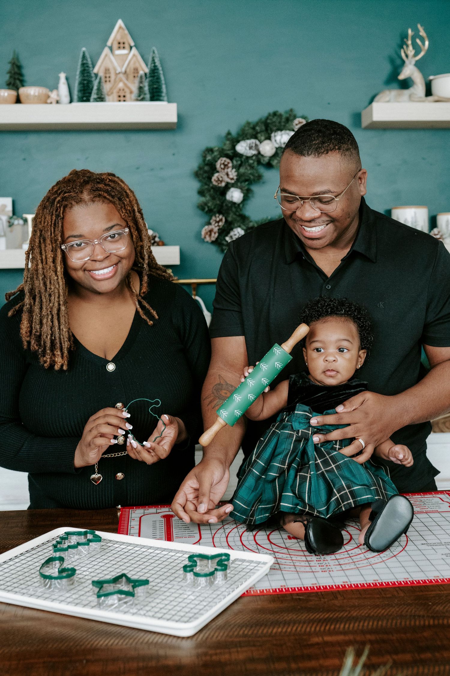 Family decorating Christmas cookies together with green cookie cutters on a white mat at wooden table.