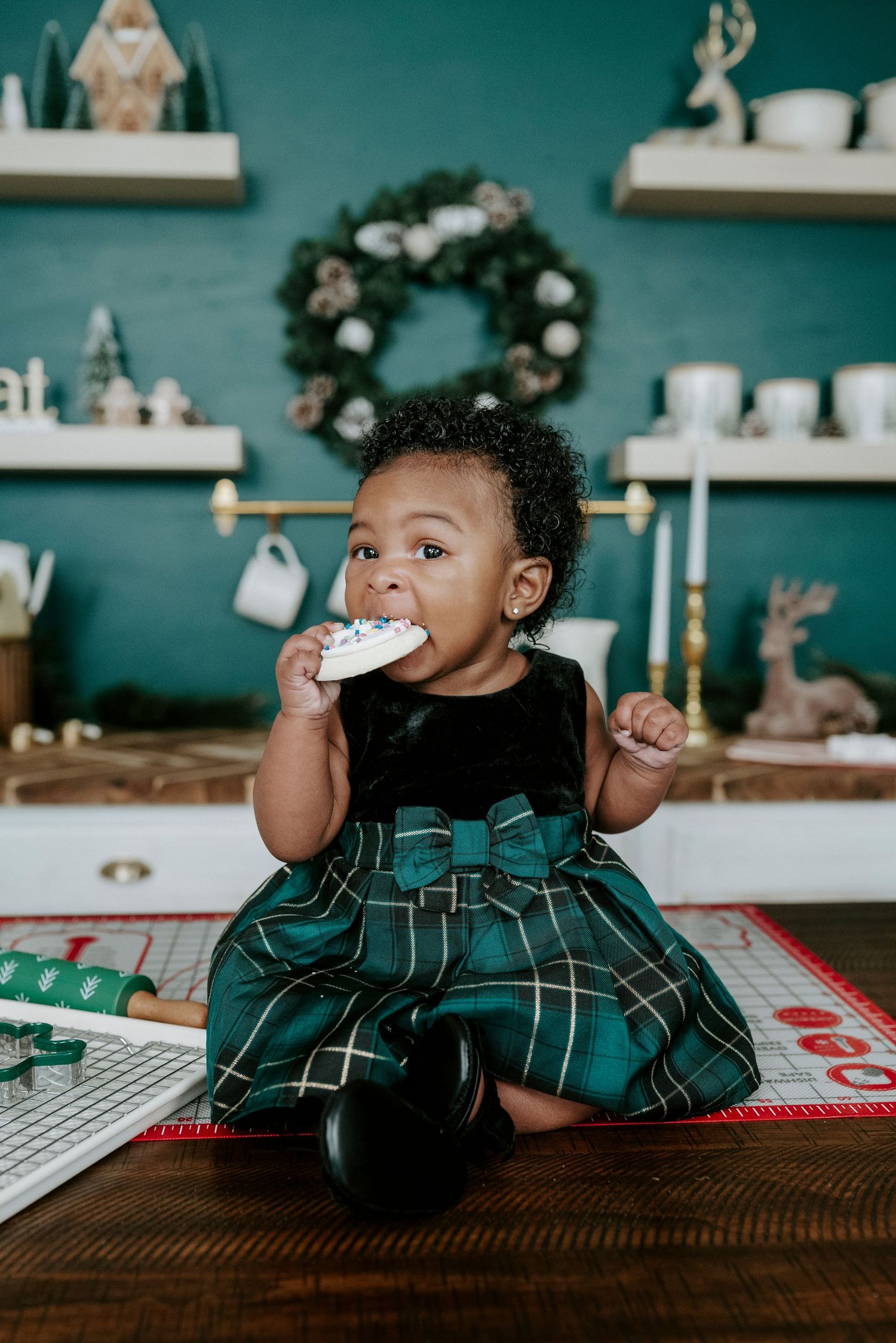 A small child in a green plaid dress enjoys a holiday treat while sitting on a table with festive decor in the background.