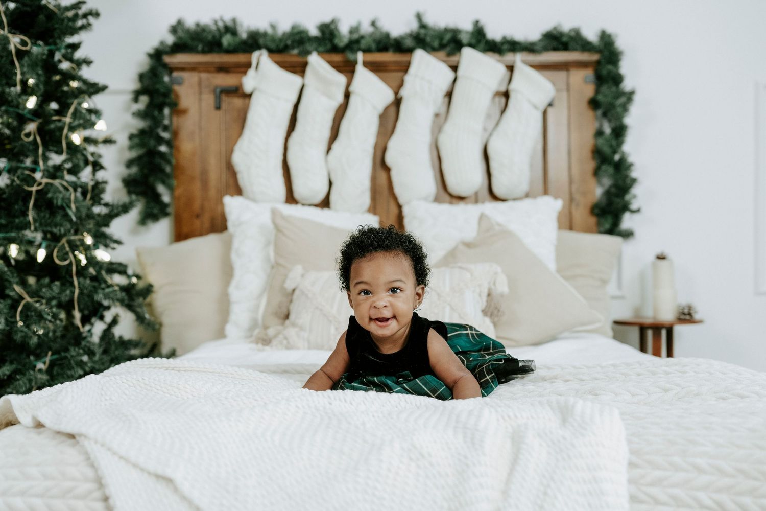 A small child in a dark dress lies on a white bed with Christmas decor and stockings hanging on the headboard.