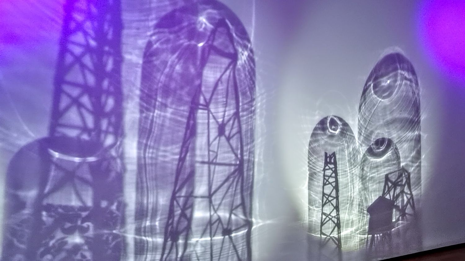 Purple and blue illuminated industrial tower silhouettes projected as shadows on a white wall.