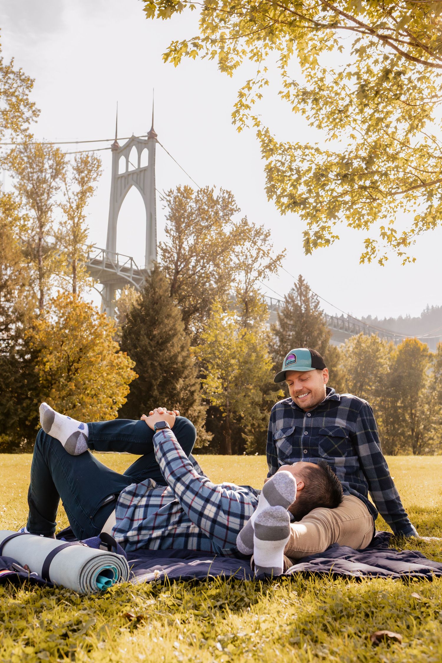 Two people relax on a blanket in a park with St. Johns Bridge and autumn foliage in Portland, Oregon.