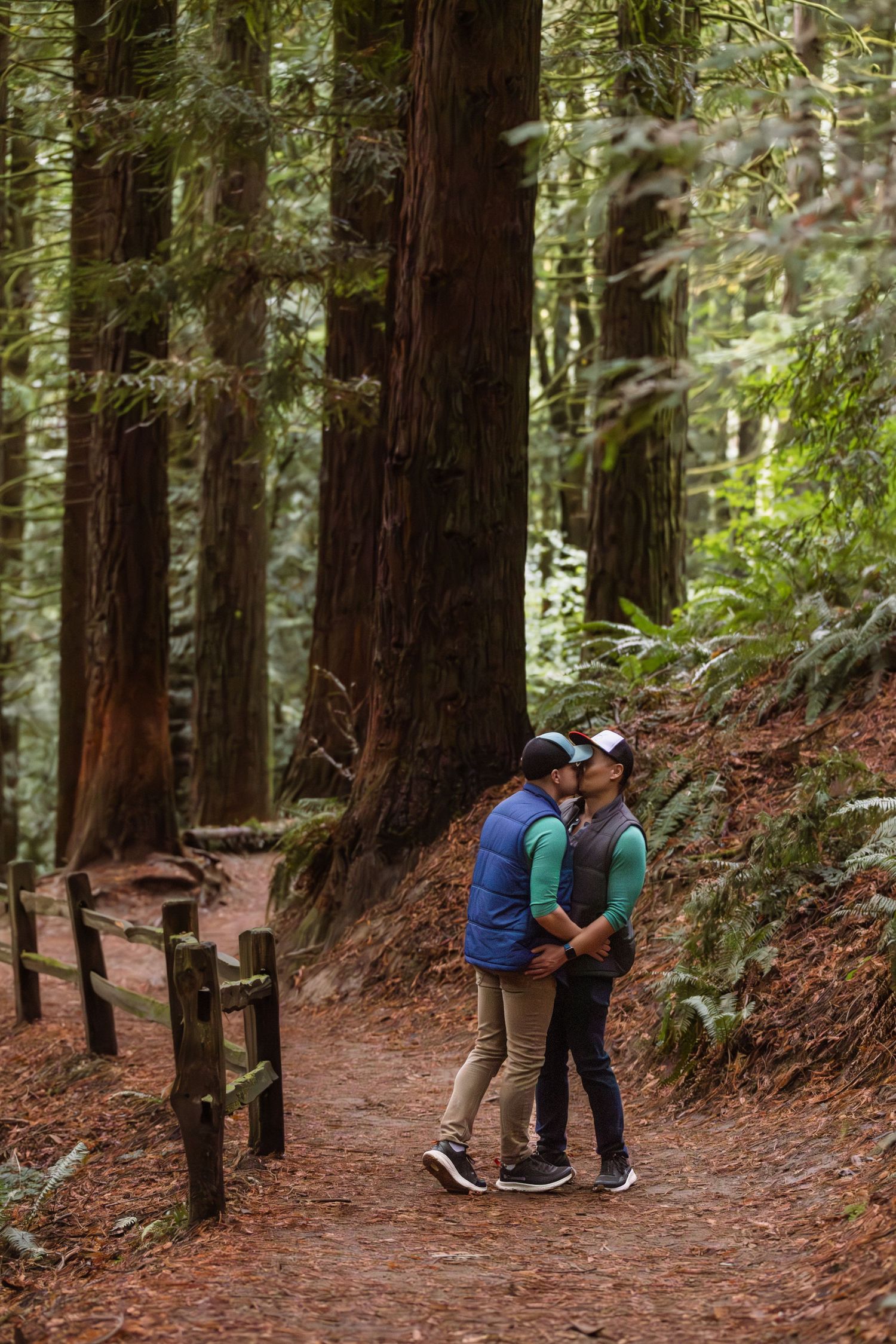 Two people share an intimate moment on a trail surrounded by towering redwood trees in a misty forest setting.